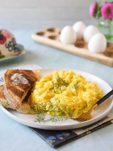 Angled view of scrambled eggs on a white plate with feta and dill, with eggs in the background