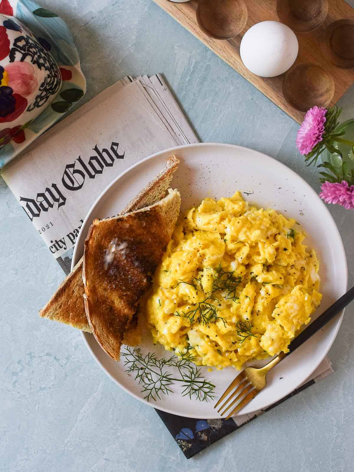 Overhead view of scrambled eggs with feta and dill with toast on a white plate with a newspaper placed underneath