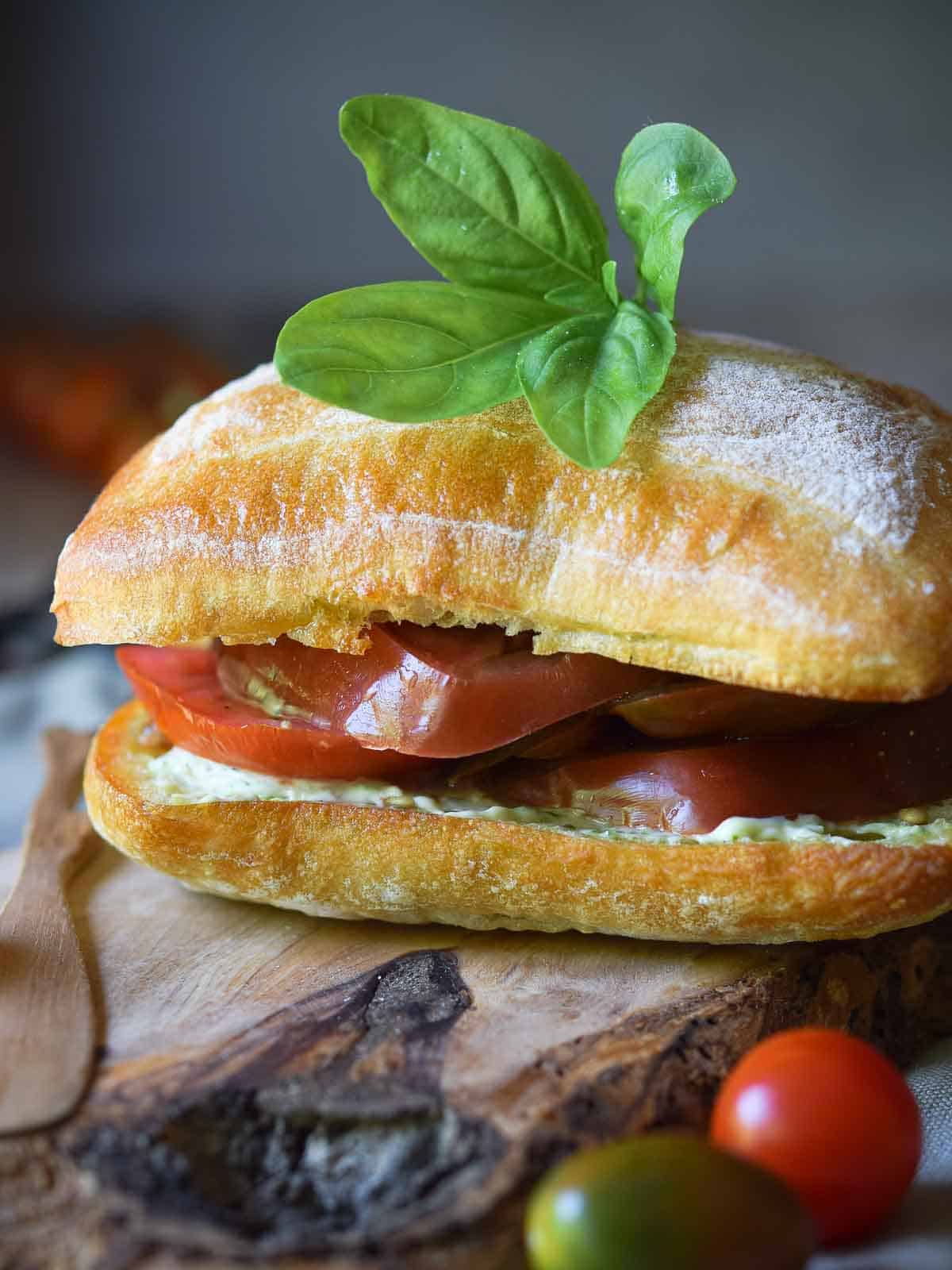 Ciabatta roll with tomatoes and pesto, topped with a few leaves of fresh basil on a wood board.