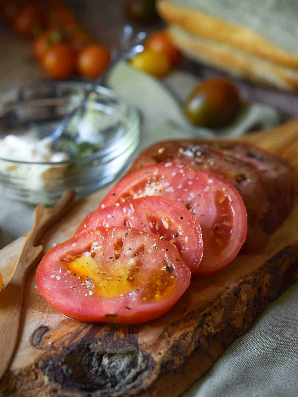 Sliced seasoned tomatoes on a wood board, with bread and pesto in the background.