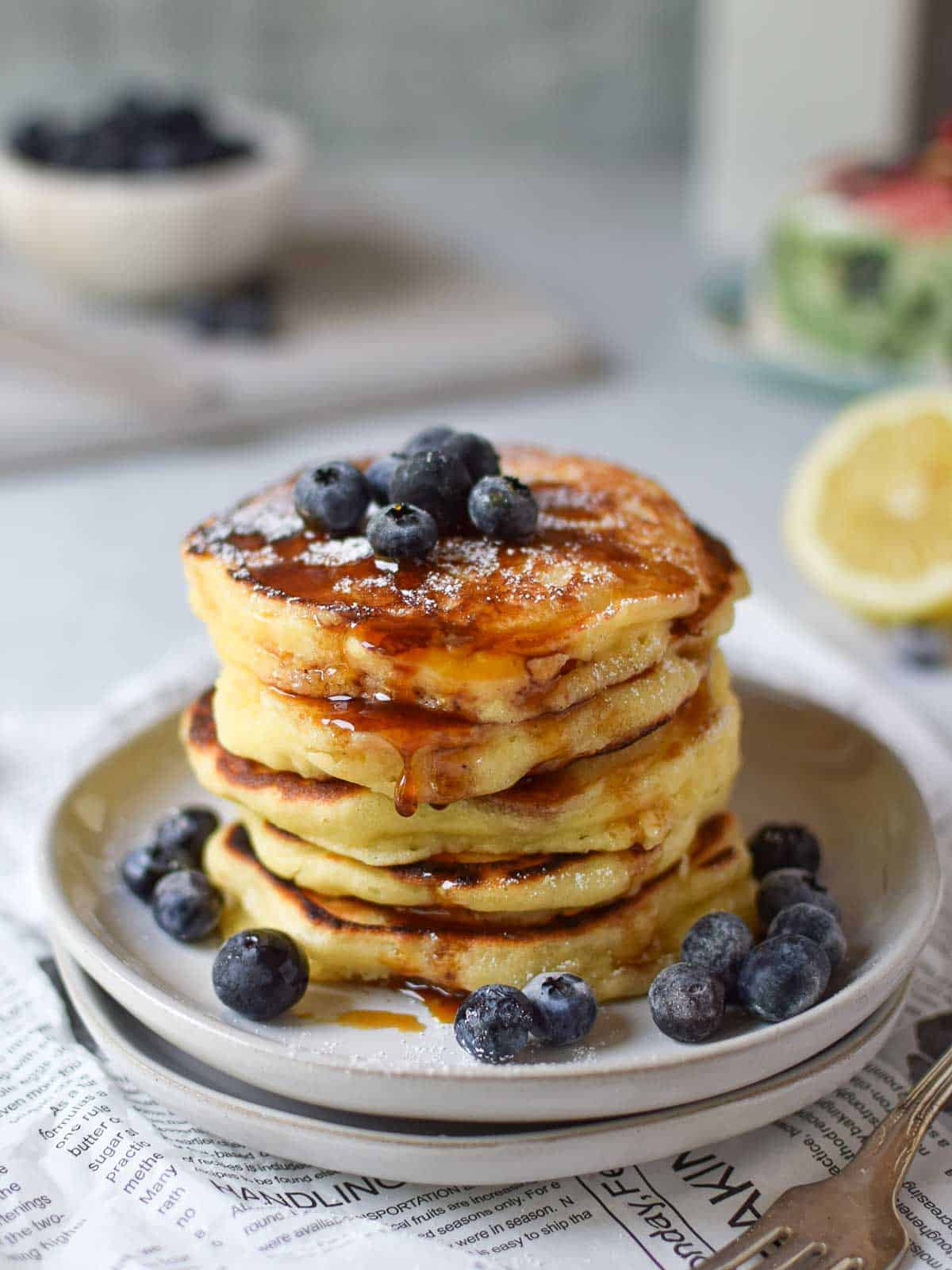 Lemon pancakes with maple syrup stacked on plates with blueberries on top.