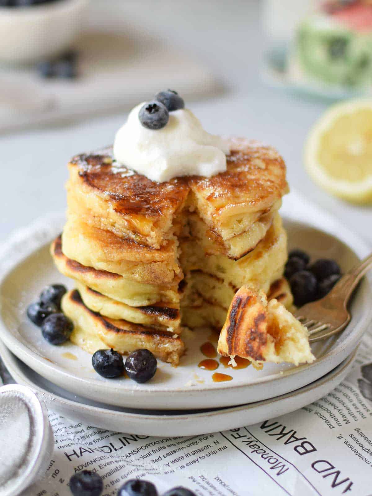Lemon mascarpone pancakes stacked on plates with blueberries on top and bites taken of them.