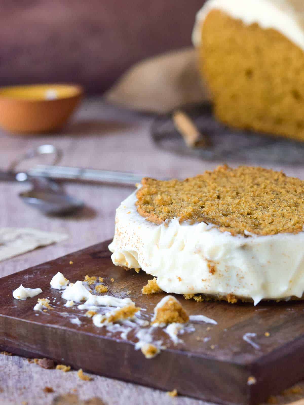 Close-up of a slice of pumpkin bread with frosting with crumbs on a wood board.