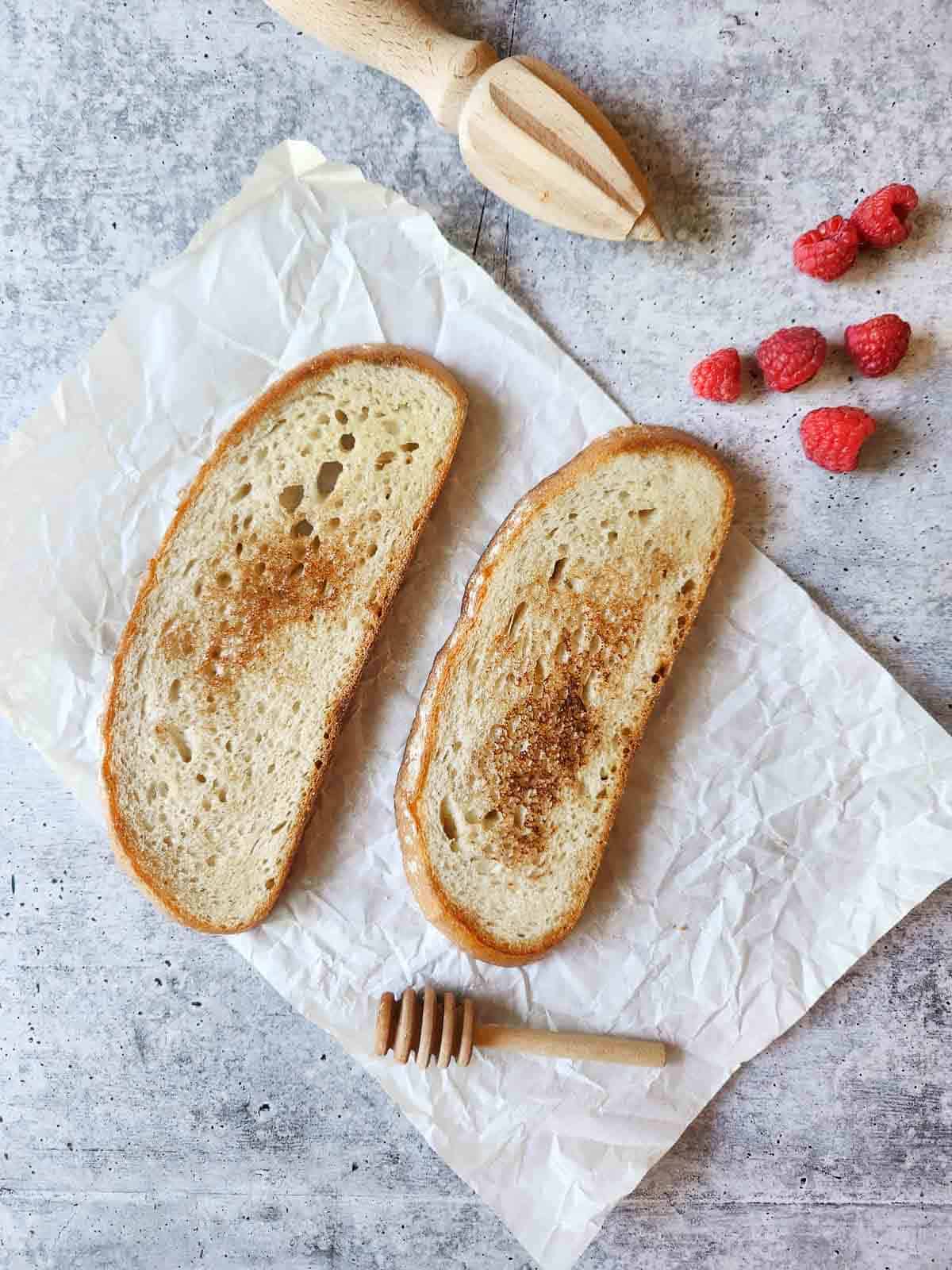 Two slices of toast on parchment paper with raspberries on the side.