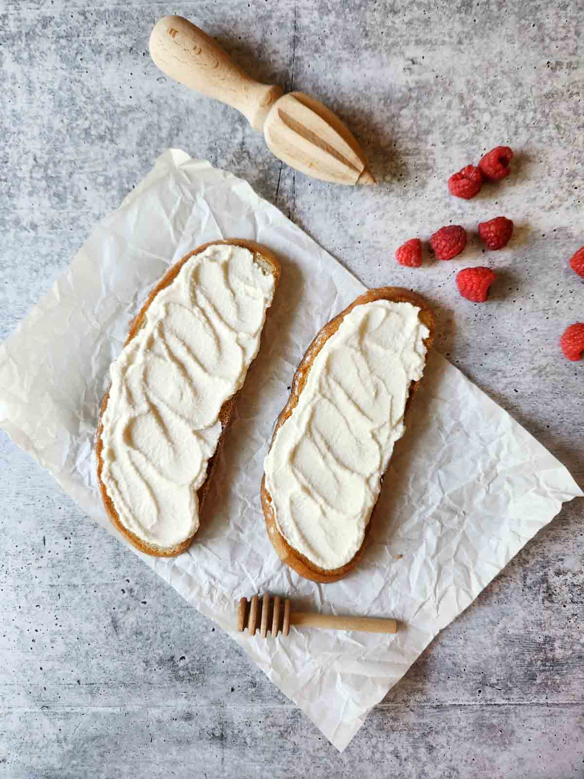 Two slices of toast with ricotta spread on parchment paper with raspberries on the side.