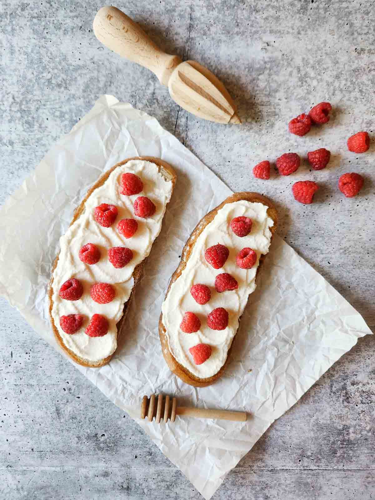 Two slices of toast with ricotta, and raspberries on white parchment paper.