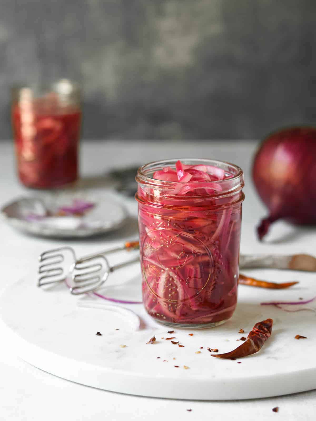Red-onions in a jar with spices around it on a marble surface.