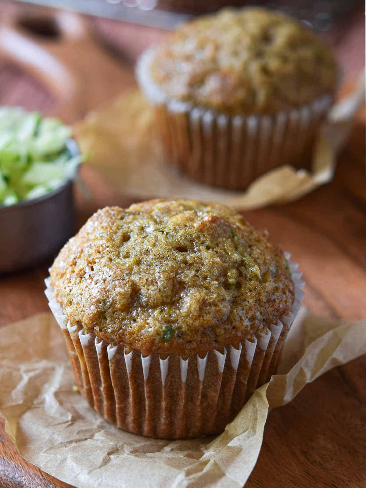 Two muffins on a wood surface with shredded zucchini in the background.