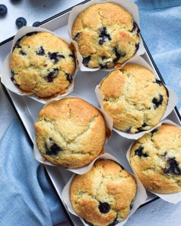 Overhead view of blueberry lemon muffins in a white tin, on a white background with a blue napkin next to it.