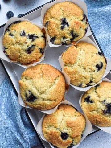 Overhead view of blueberry lemon muffins in a white tin, on a white background with a blue napkin next to it.