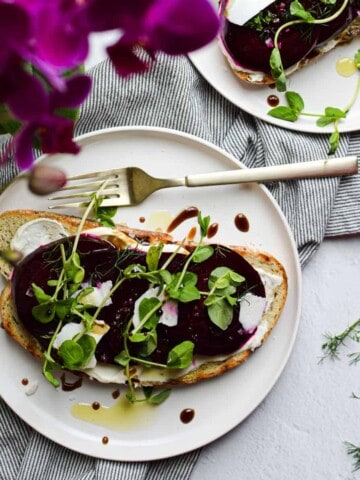 Overhead view of toast with sliced roasted beets, creme fraiche and dill on a whote plate with bright pink flowers next to it