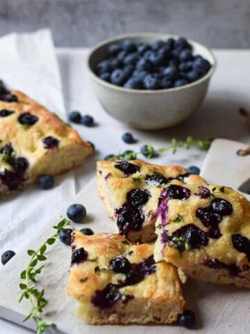 Angled view of blueberry and thyme focaccia on a serving grey serving board with blueberries in the background.