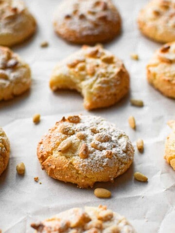 Angled view of baked Italian pignoli cookies on a baking sheet covered with white parchment paper. One cookie has a bite taken out.