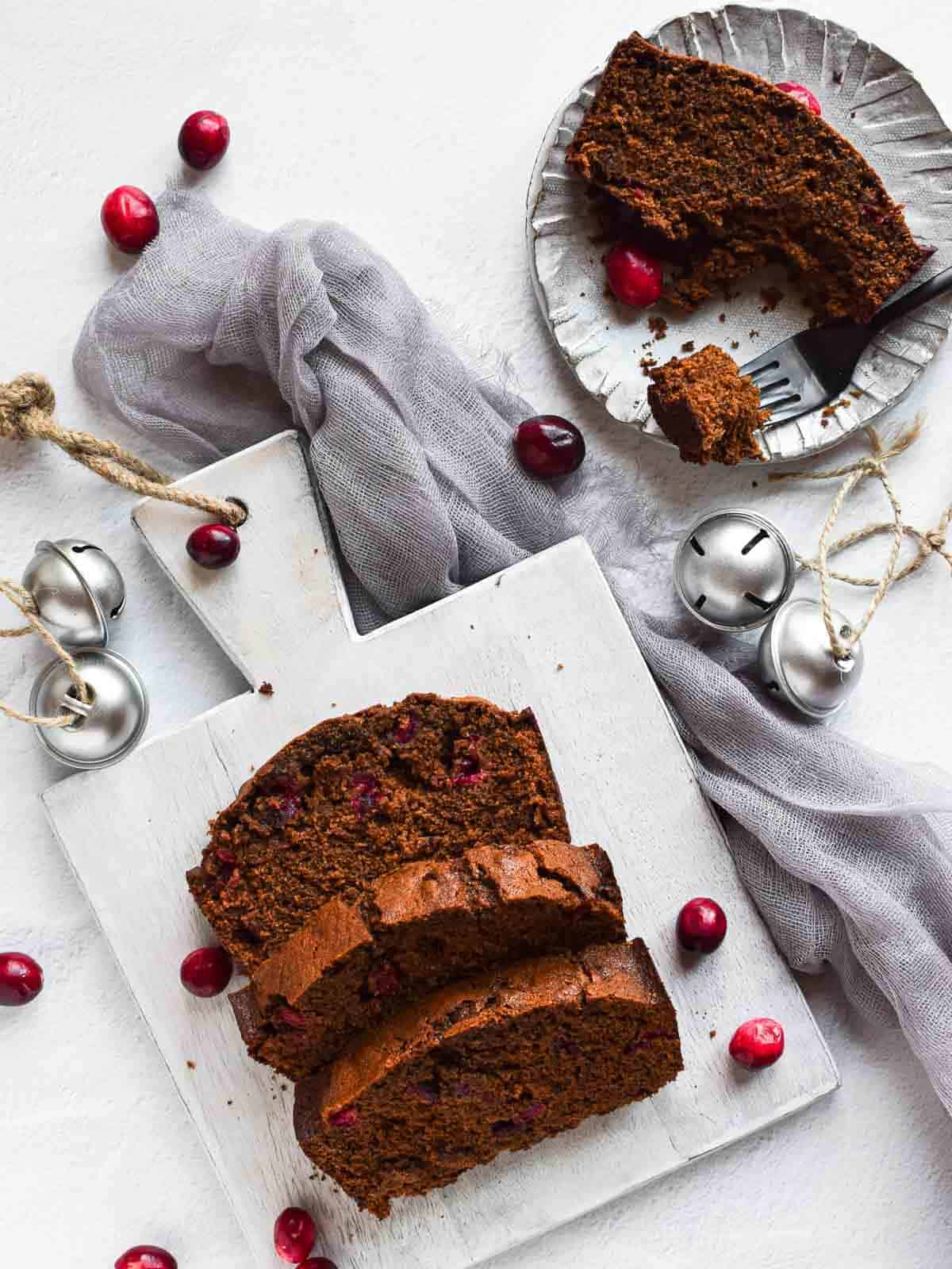 Overhead view of cranberry gingerbread loaf on a grey cutting board, with slice on a plate on a white background.