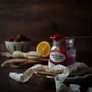 Eye-Level view of strawberry rhubarb jam in a jar against a brown background with strawberries in the distance.