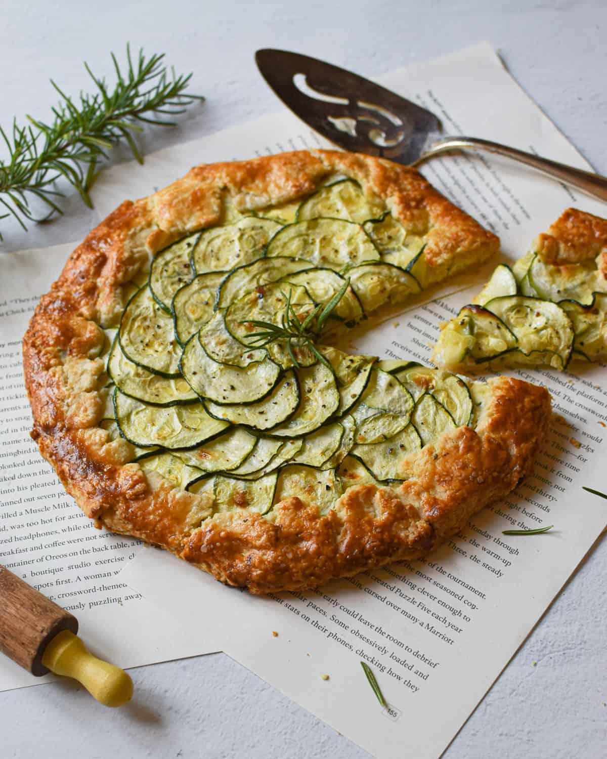 Angled view of zucchini ricotta galette with a slice cut out surrounded by scattered rosemary on a white background.
