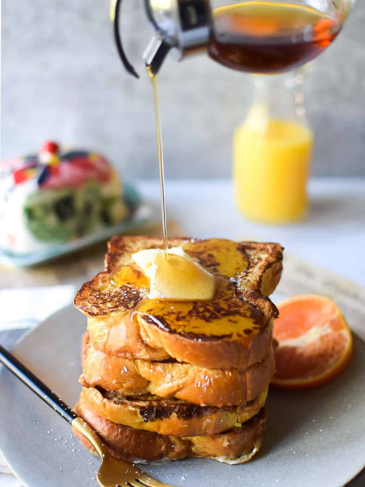 Straight on view of orange and cardamom french toast with maple syrup being poured from above on a grey background