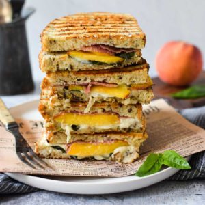 Eye level view of peach, prosciutto and burrata panini stacked high on a white plate with a peah in the background.