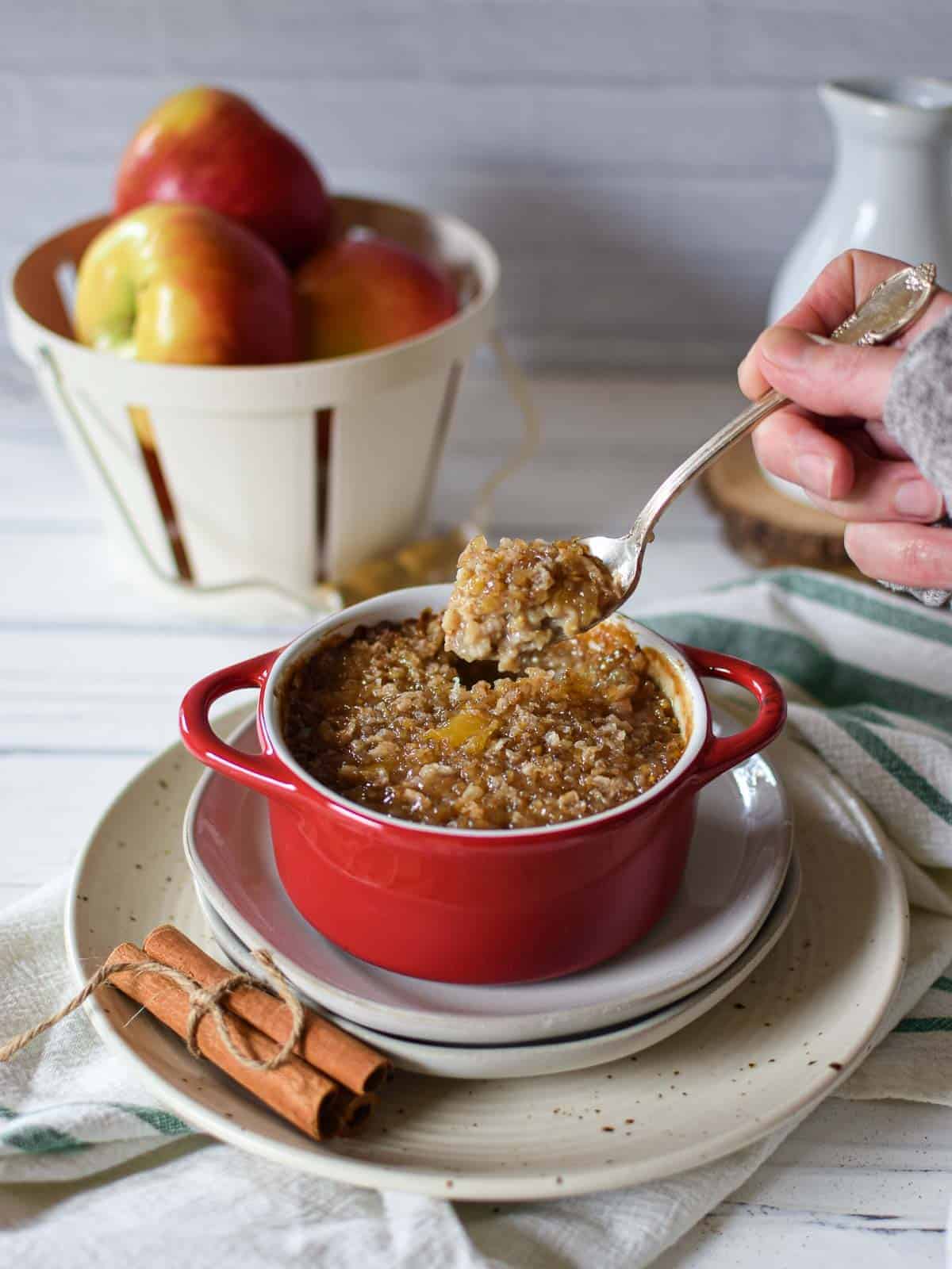 Angled view of apple pie brulee oatmeal in a red dish with a hand scooping out a bite, on a white background with apples in the background