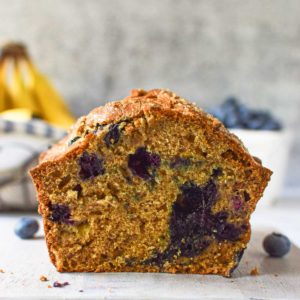 Eye-level view of roasted banana blueberry bread cut in half on a white background with a bunch of bananas in the background