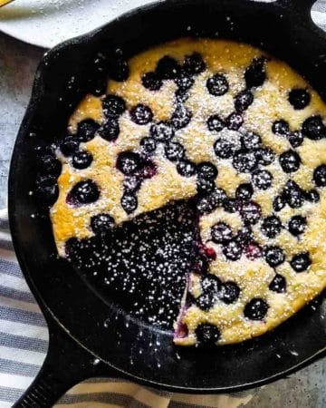 Overhead view of blueberry pancake skillet with a slice cut out on a plate in a black skillet