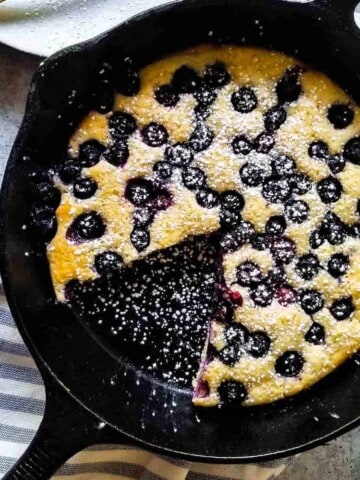 Overhead view of blueberry pancake skillet with a slice cut out on a plate in a black skillet