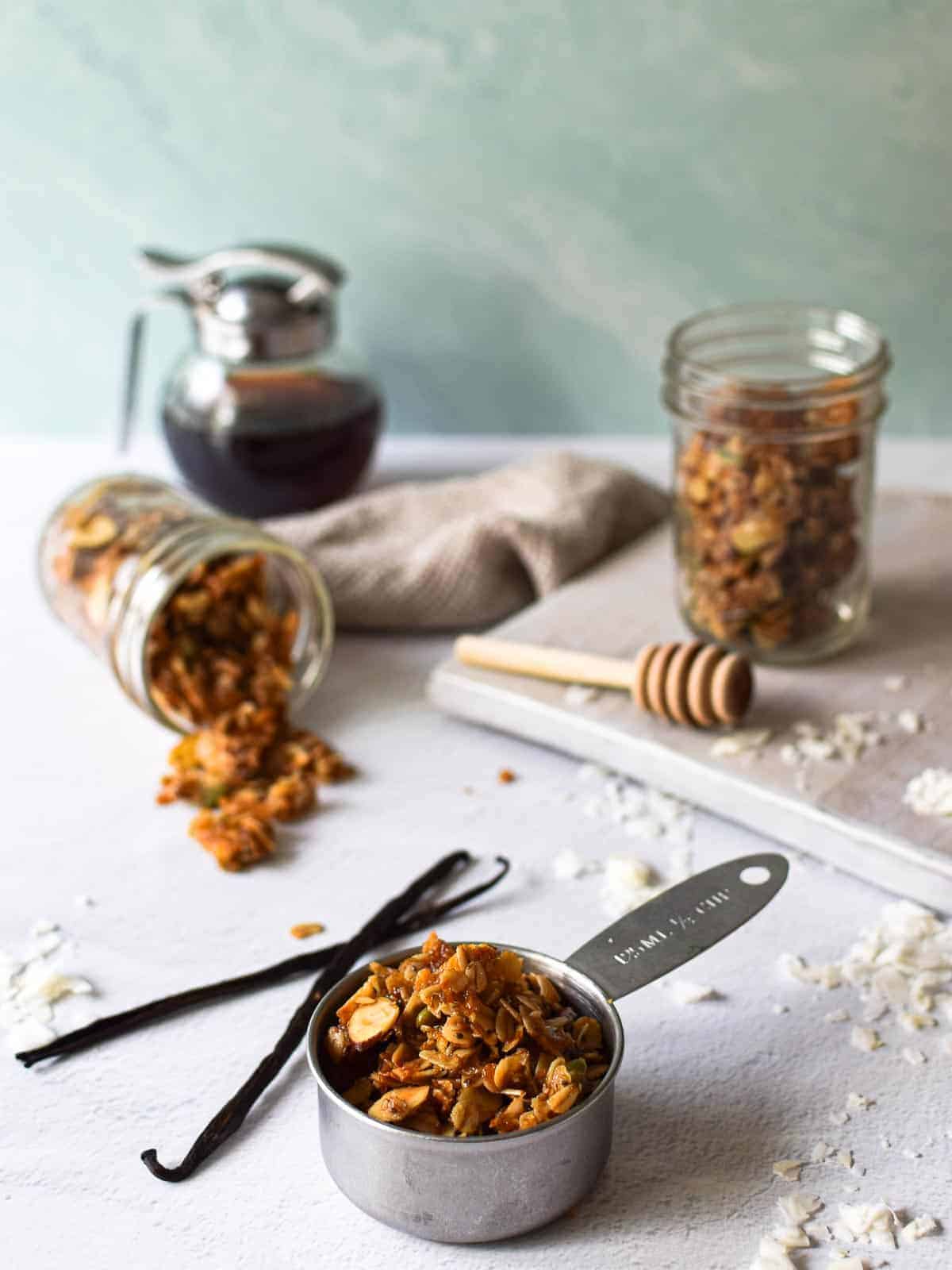 Angled view of vanilla coconut granola in jars and a measuring cup with vanilla bean and coconut scattered