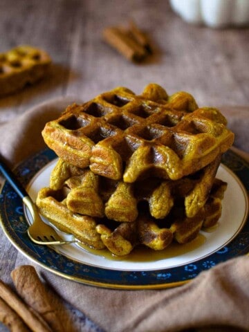 Pumpkin waffles stacked on a plate with maple syrup and cinnamon on a wooden surface.