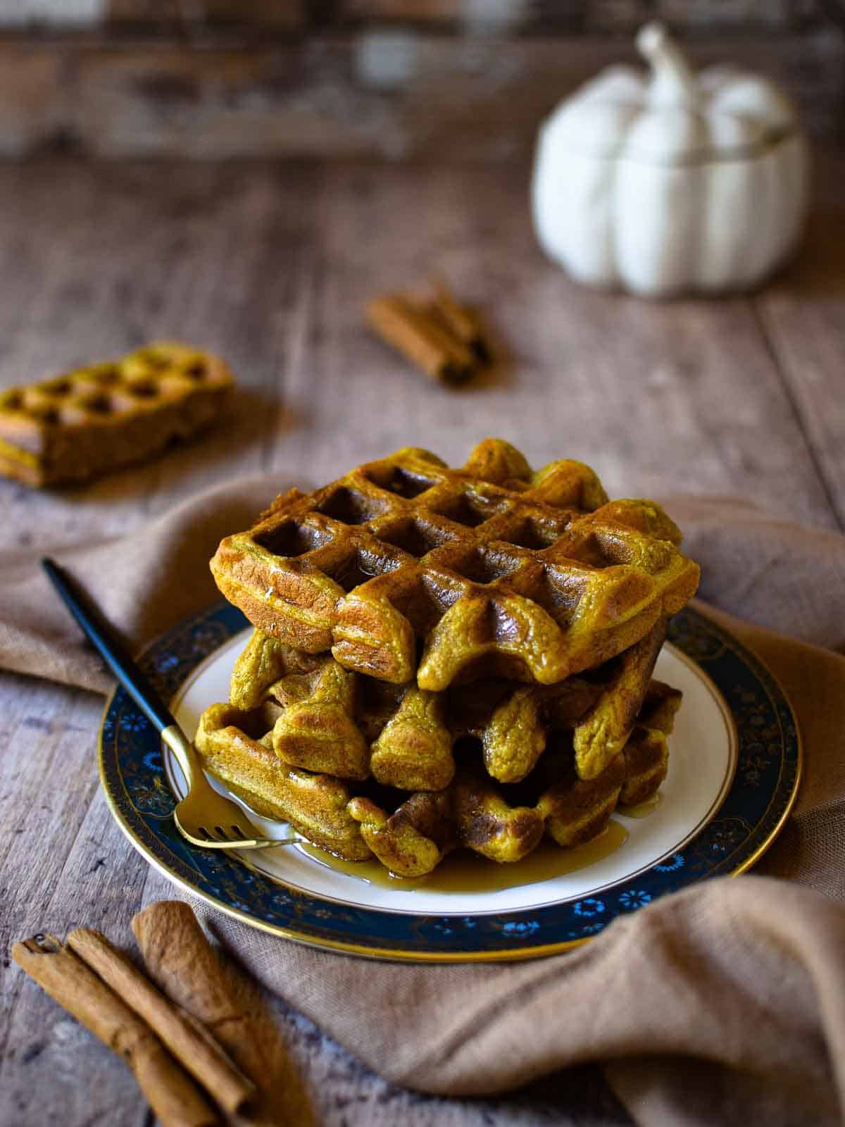 Pumpkin waffles stacked on a plate with maple syrup and cinnamon on a wooden surface.