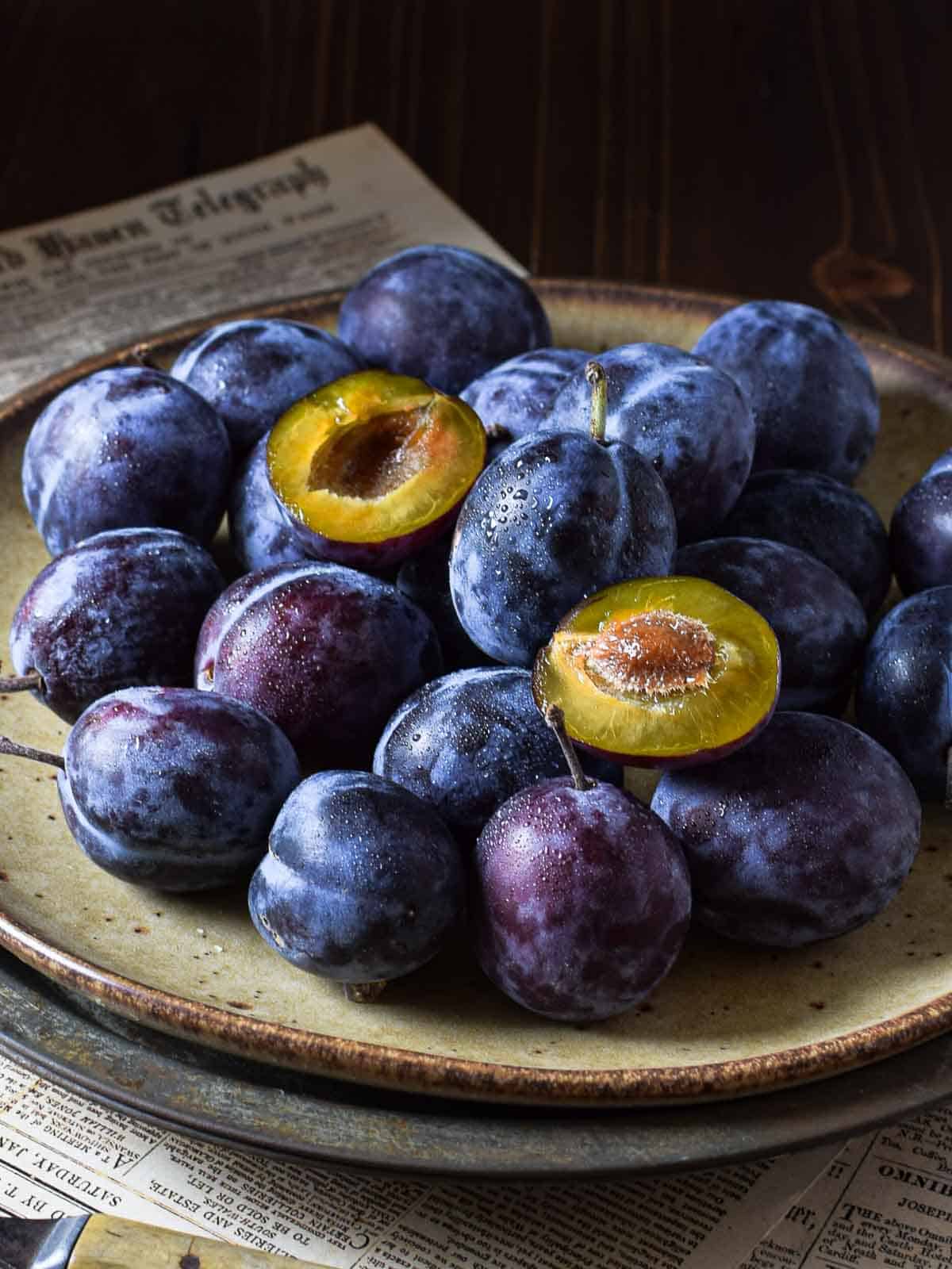 Italian plums stacked on a brown plate with 2 plums cut open.