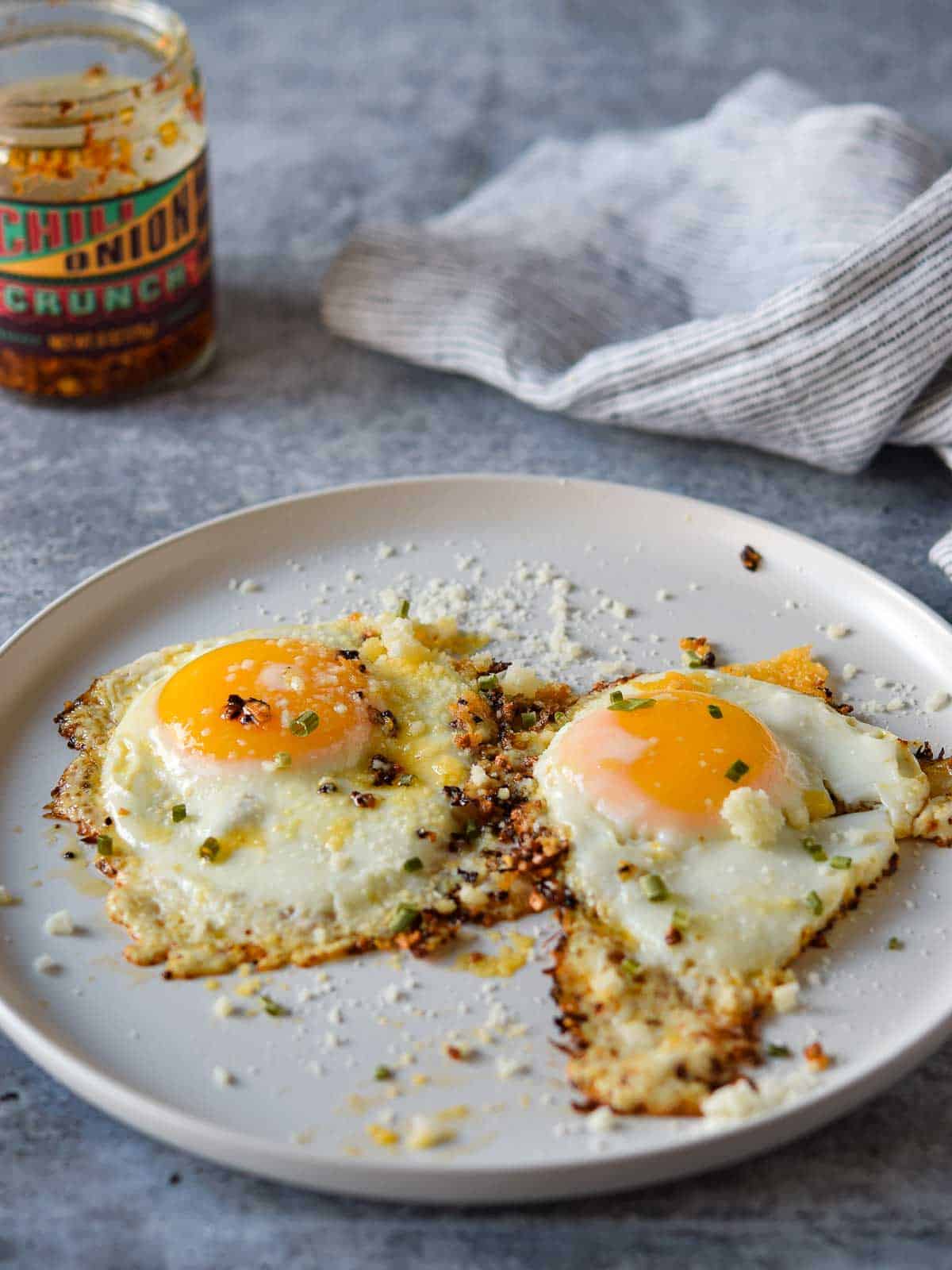 Angled view of two fried eggs on a plate with a jar of chili crunch in the background.
