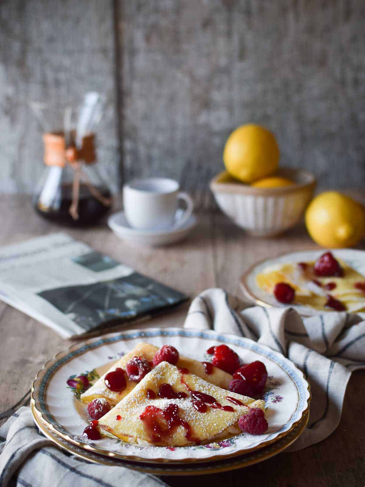 Stuffed crepes on a flowered plate with raspberry sauce on top, with coffee and lemons in the background.