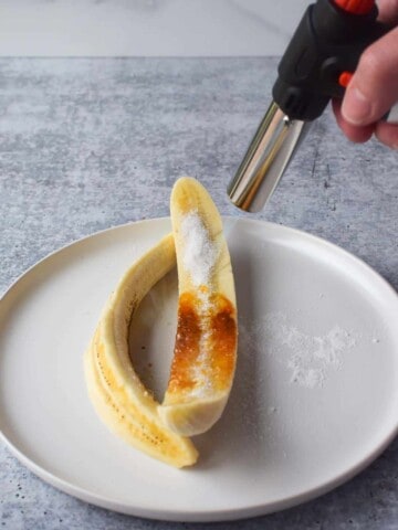 Sliced banana on a white plate getting caramelized with a culinary torch.