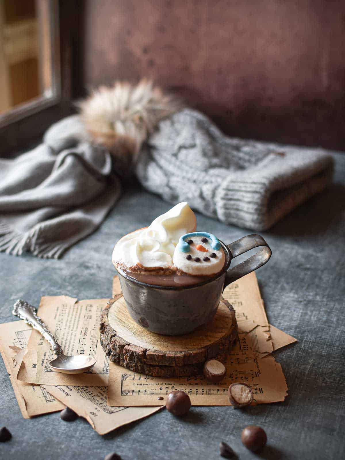 Hot chocolate scene by a window with a hat and scarf in the background and scattered malted milk balls.