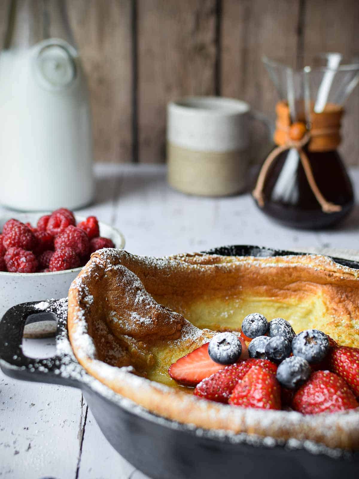 Eye-level view of a Dutch baby in a cast iron skillet with fruit and coffee in the background.