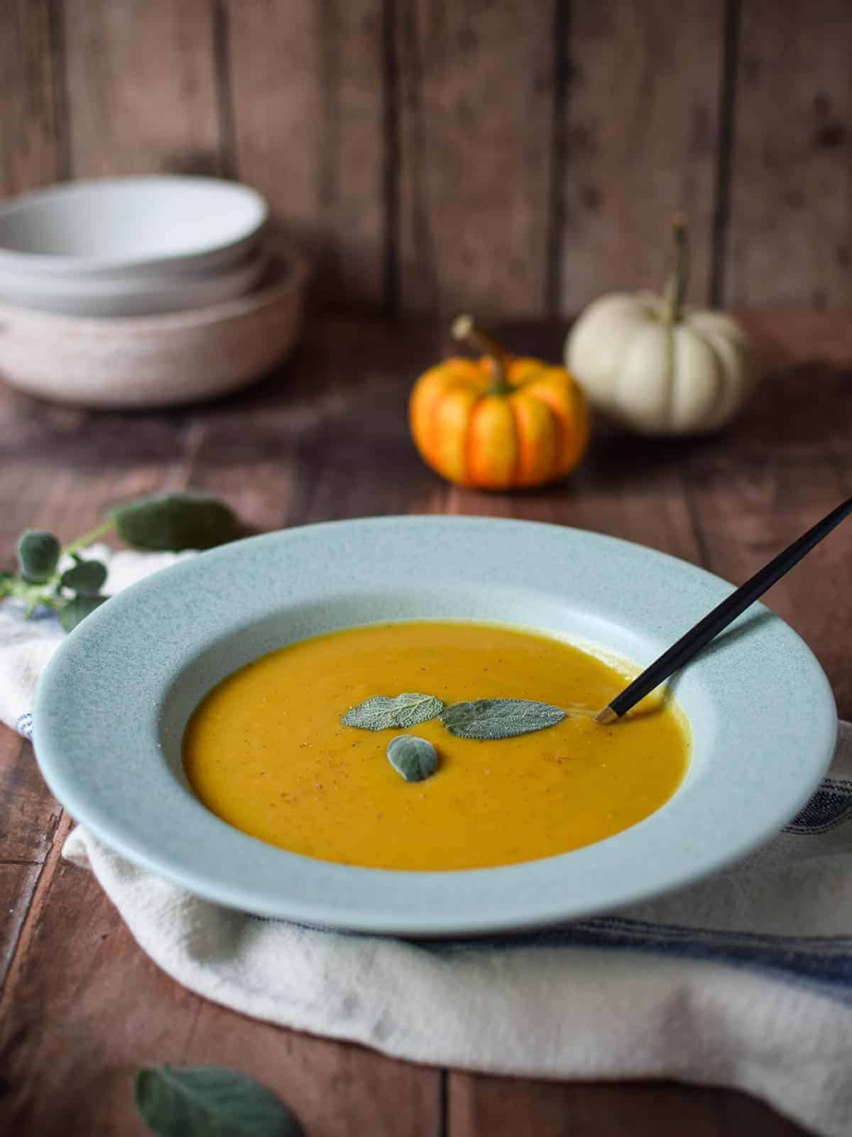 Squash soup in a blue bowl with sage and pumpkins in the background.