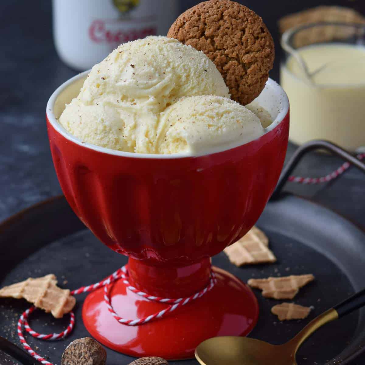 Eggnog ice cream in a red ice cream bowl with waffle cone pieces and gingersnaps around it.