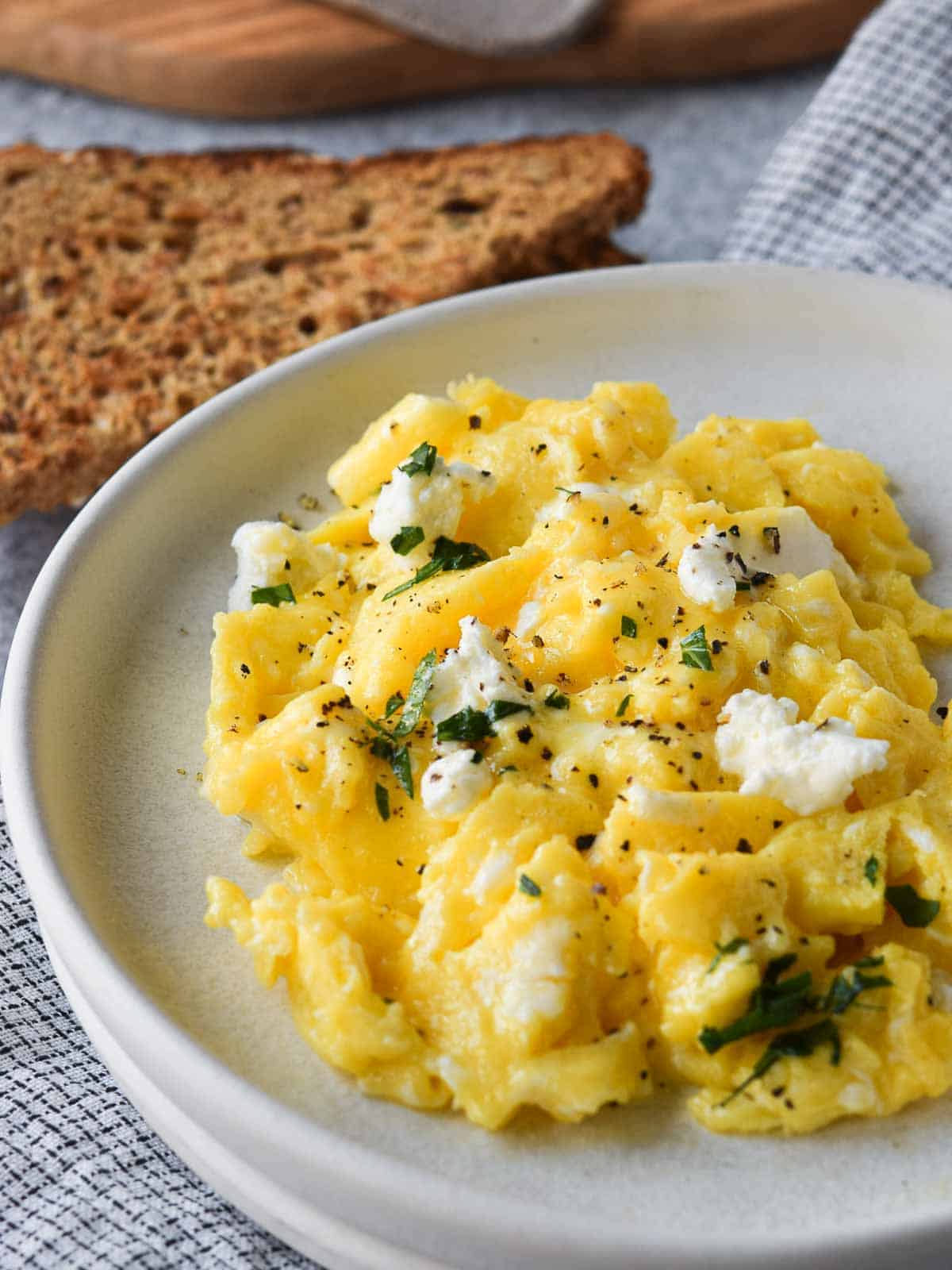 Scrambled eggs with goat cheese on a white plate with toast in the background.