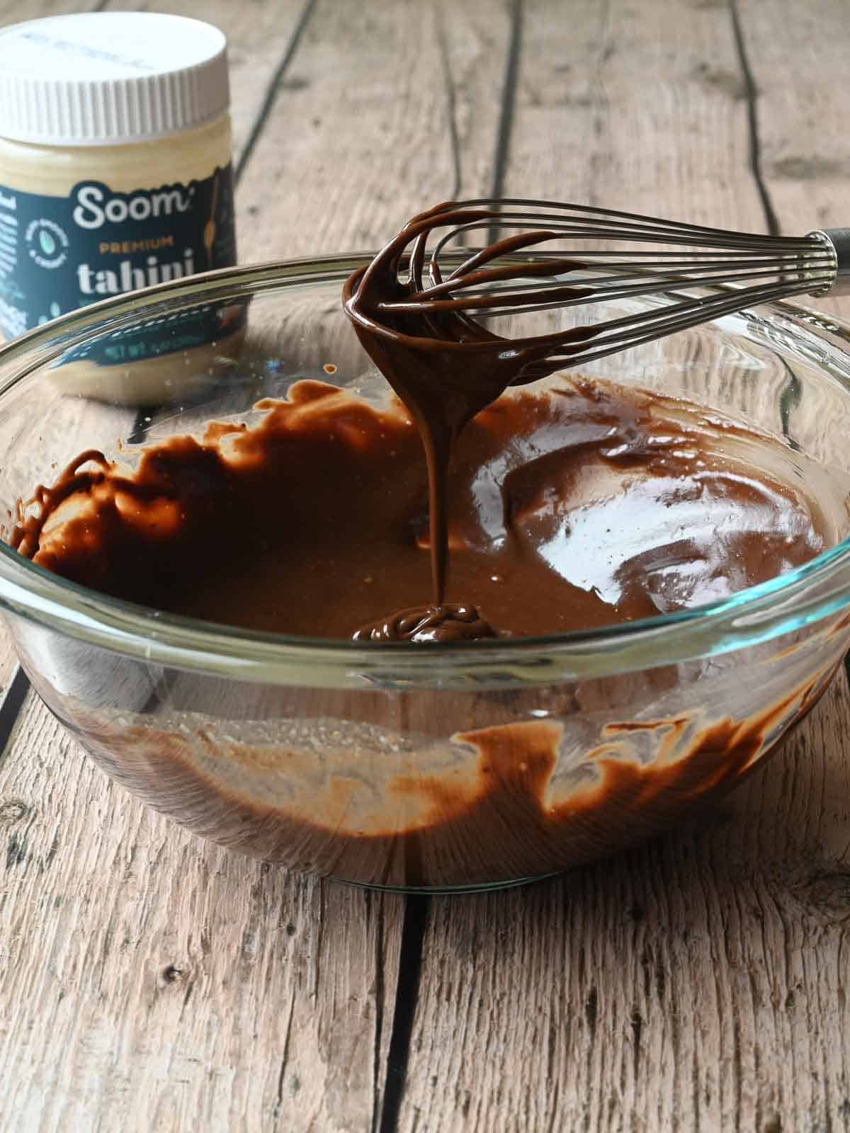 Chocolate tahini in a clear mixing bowl with a whisk on the side with dripping chocolate.