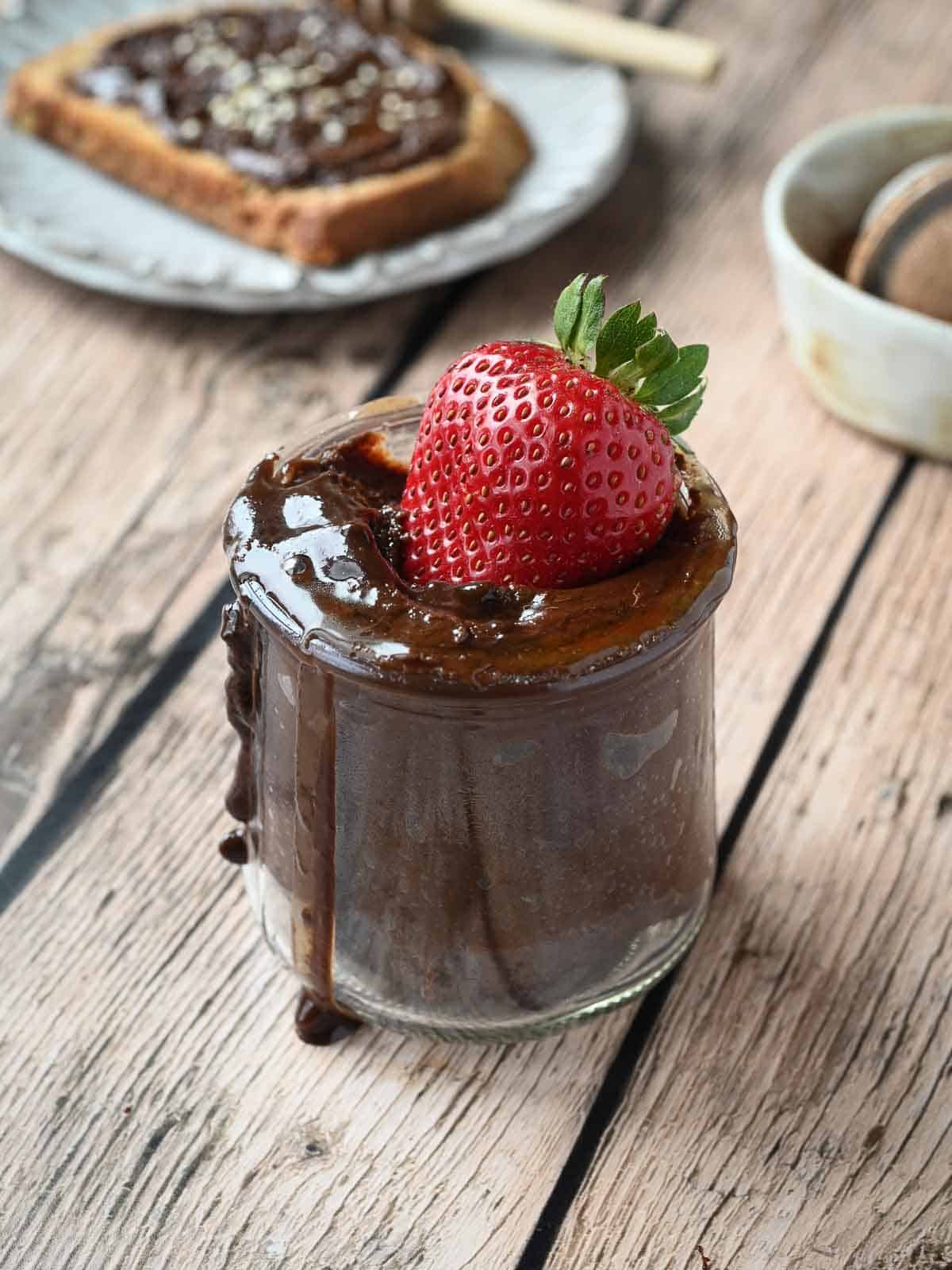 A strawberry dipped into a jar of chocolate tahini spread on wood.