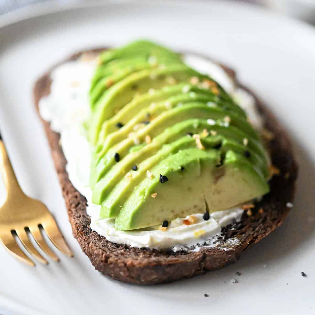 Slices of avocado and cream cheese on pumpernickel bread on a white plate.