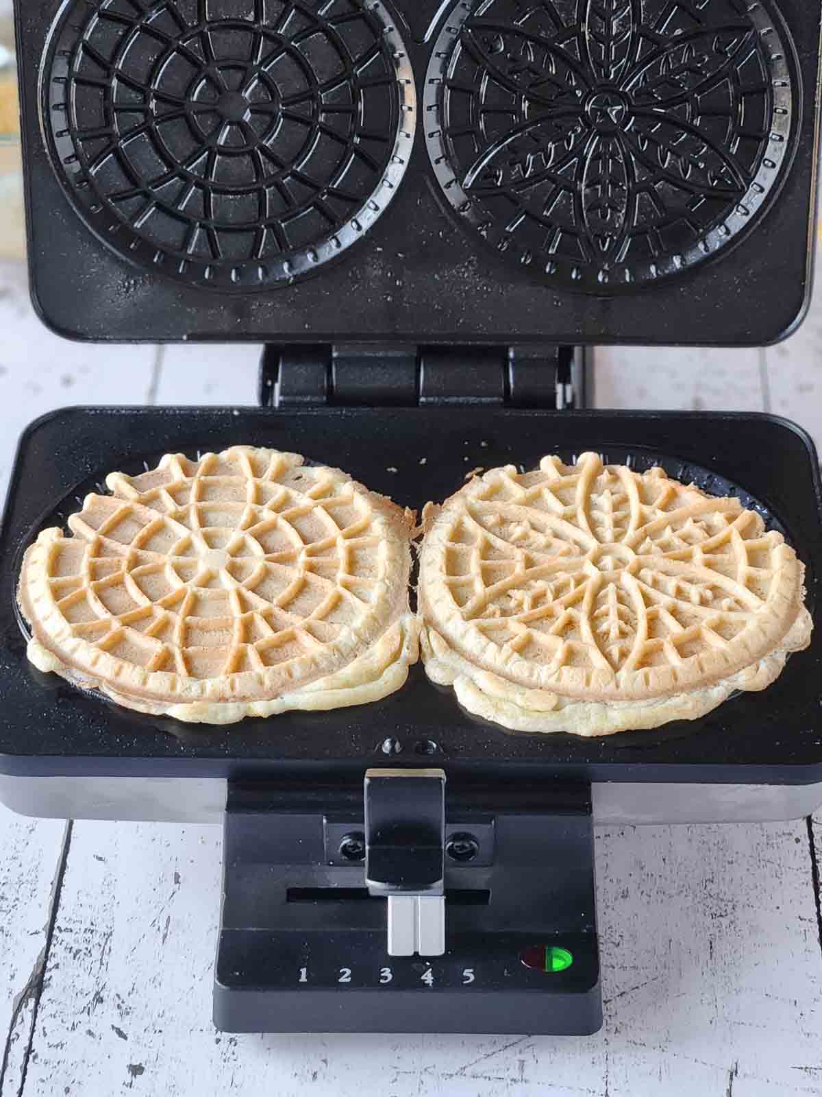 Two cooked pizzelles in a pizzelle maker with the lid open.