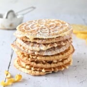 Lemon pizzelles stacked on top of each other on white wood with lemon in the background.