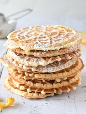 Lemon pizzelles stacked on top of each other on white wood with lemon in the background.