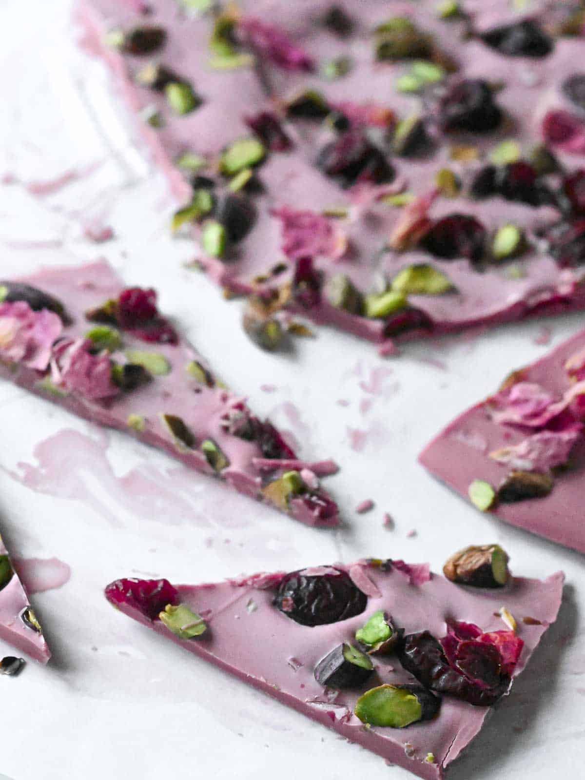 Cut up pieces of pink chocolate bark with pistachios and cranberries on white parchment paper.