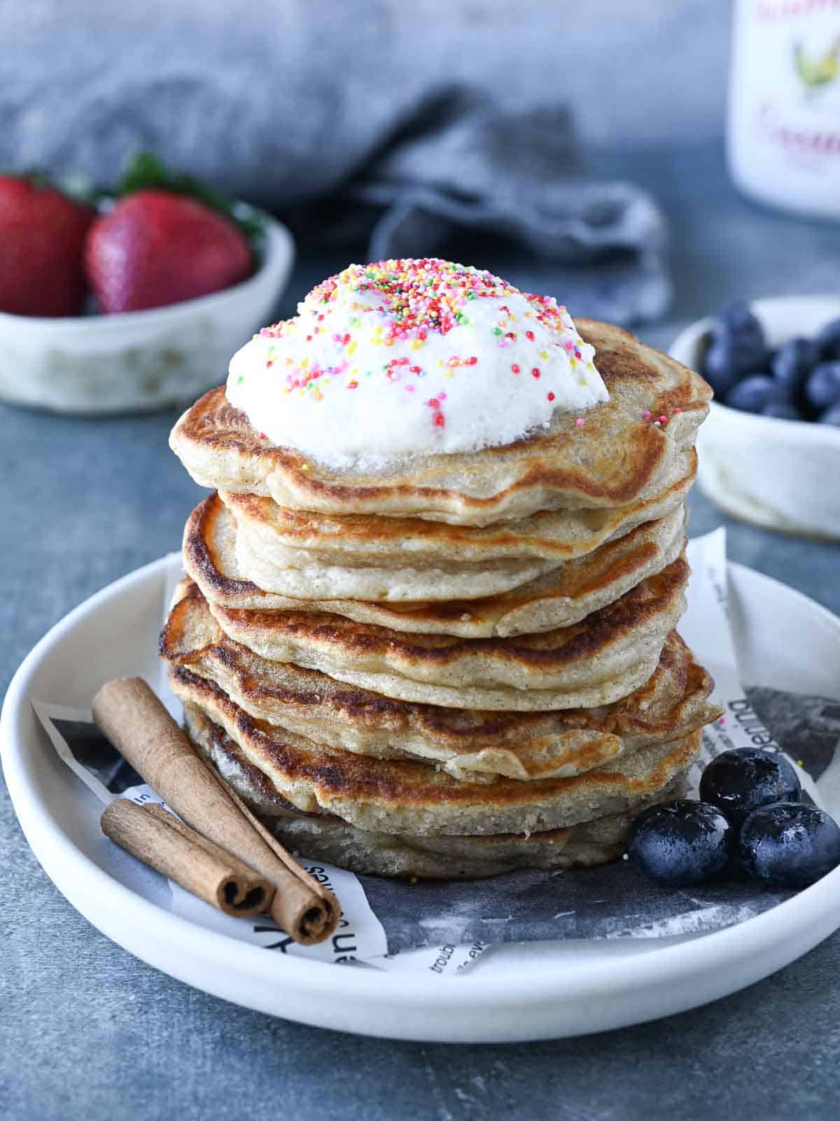 A stack of pancakes with whipped cream and sprinkles on top on a grey surface.
