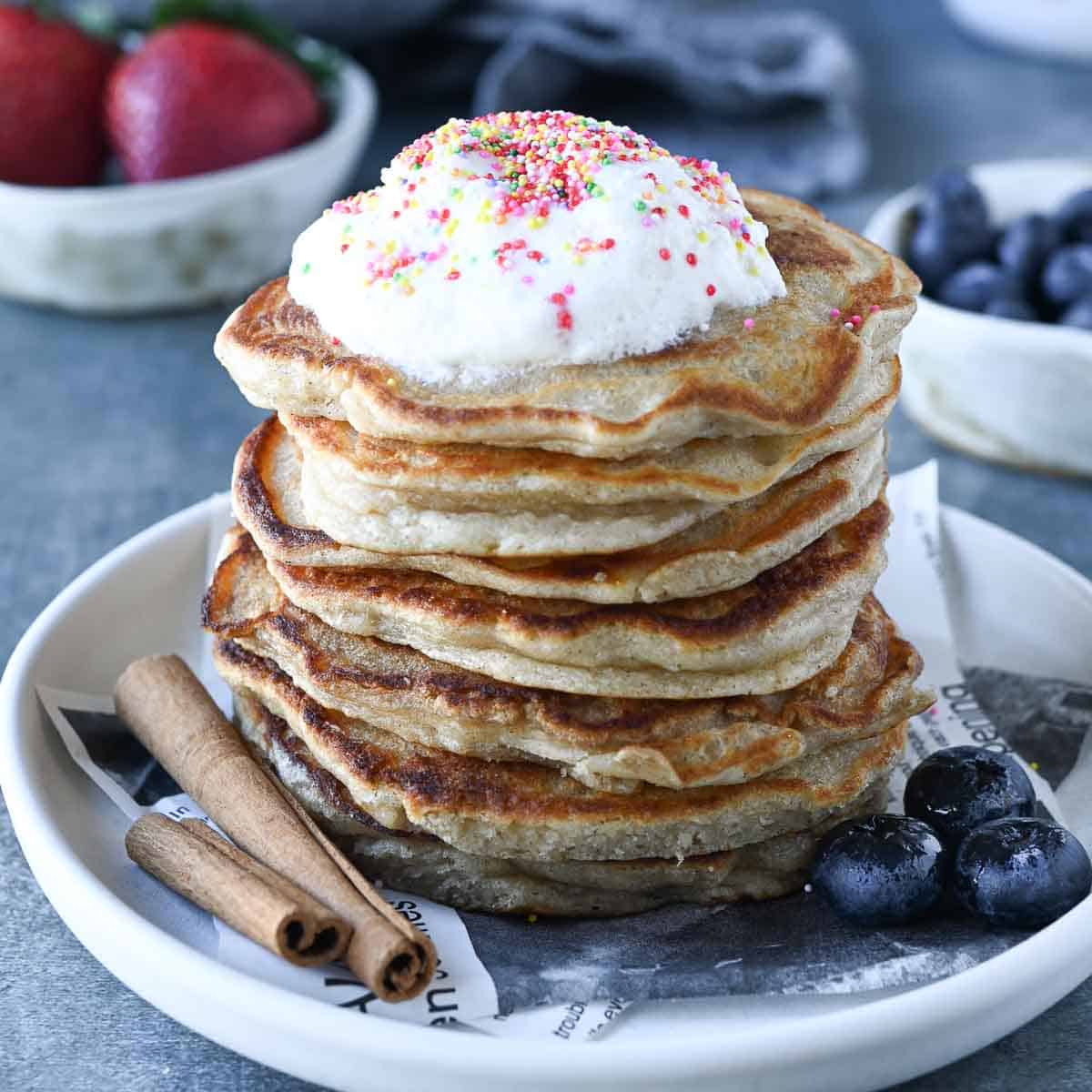 Pancakes stacked on a white plate with whipped cream, cinnamon and blueberries.