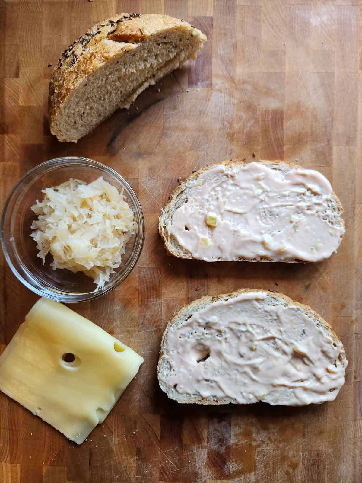 Bread, sauerkraut, dressing, and cheese on a cutting board.