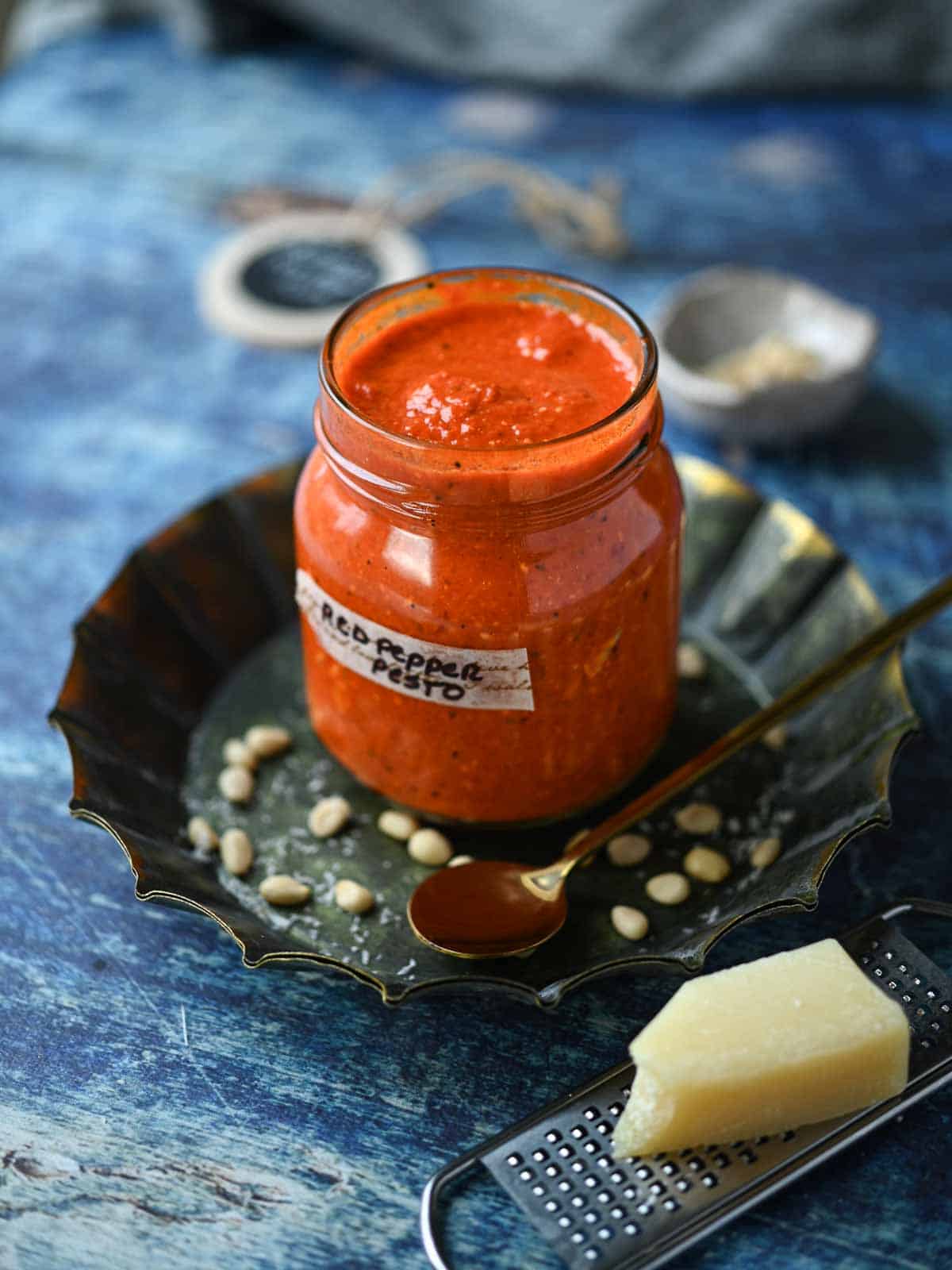 Red pepper pesto in a jar with grated cheese in the front on a blue surface.