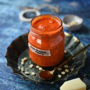 Red pepper pesto in a jar with grated cheese in the front on a blue surface.